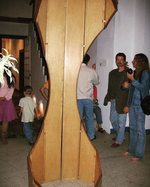 Pessoas em pé ao redor de uma escultura móvel alta de madeira clara em um espaço de galeria.
