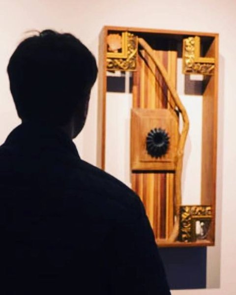 Uma figura de costas observa uma caixa de madeira vertical com relevos e molduras douradas em uma parede clara.