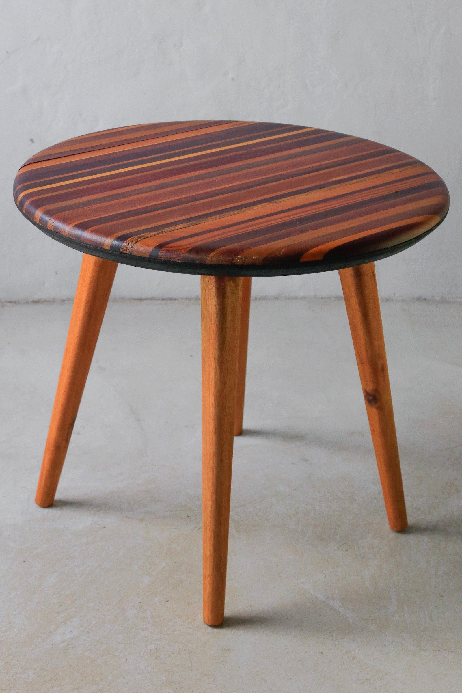 Mesa Flor com tampo circular listrado e pernas torneadas diante de parede clara.