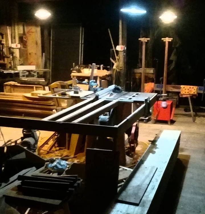 Oficina de marcenaria à noite com serras e ferramentas sob luz de lâmpadas suspensas.