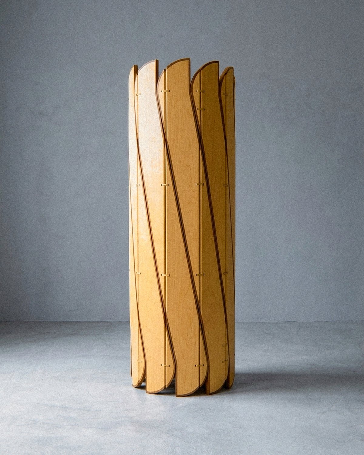 A escultura móvel Onda em compensado de maple, freixo maciço e latão fica fechada em forma cilíndrica sobre piso cinza.