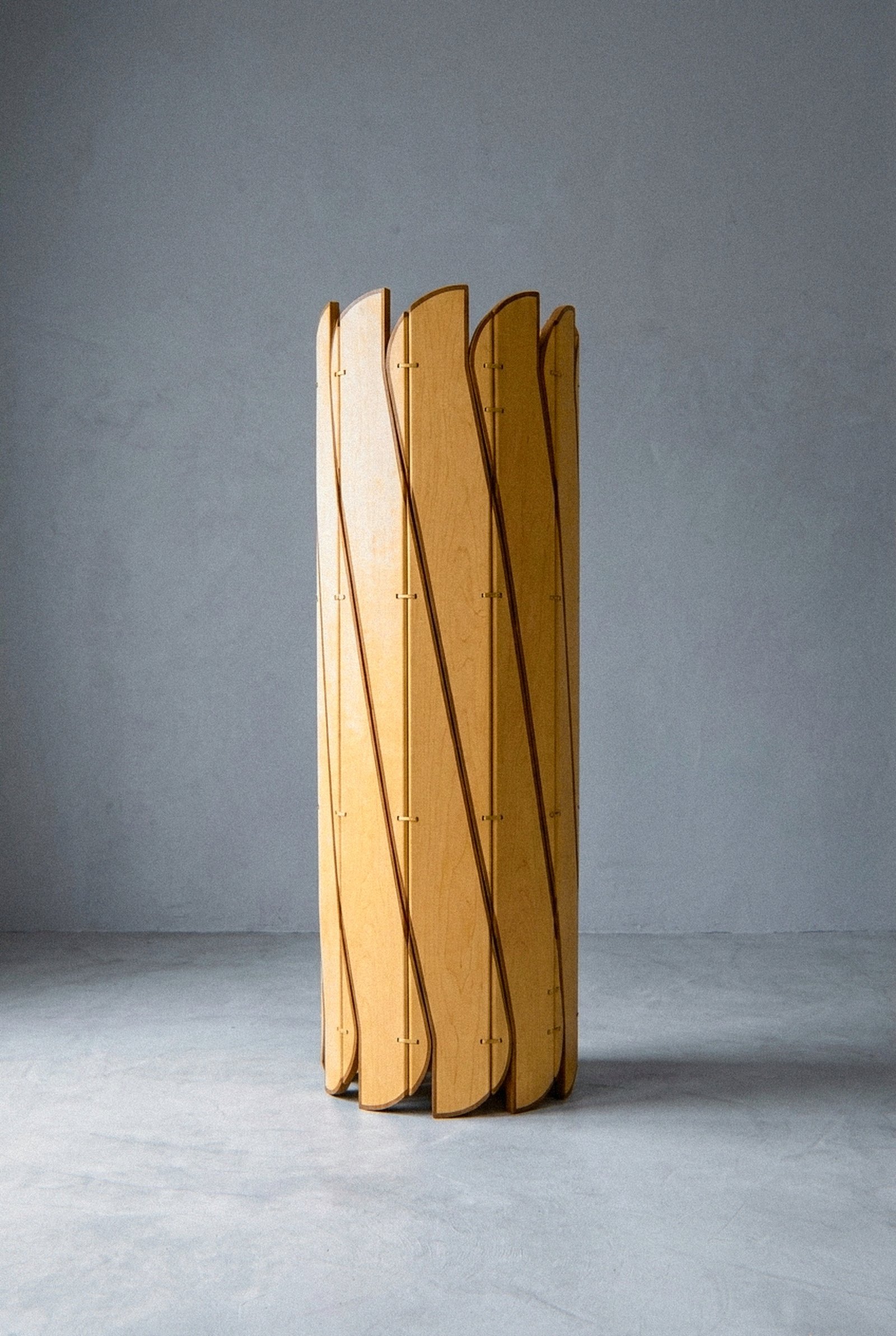 A escultura móvel Onda em compensado de maple, freixo maciço e latão fica fechada em forma cilíndrica sobre piso cinza.